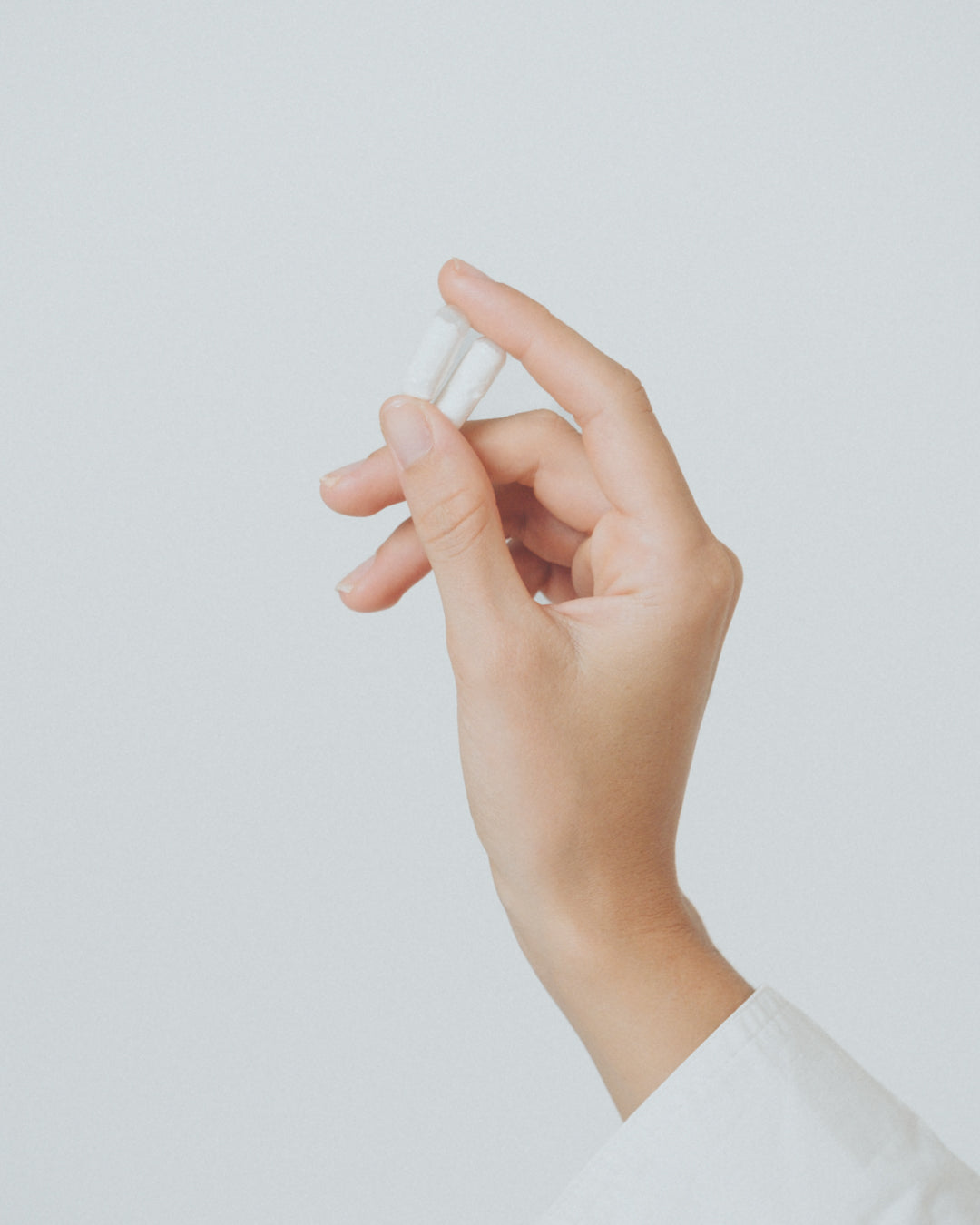 Hand hält zwei weiße Kapseln zwischen Daumen und Zeigefinger vor einem neutralen Hintergrund