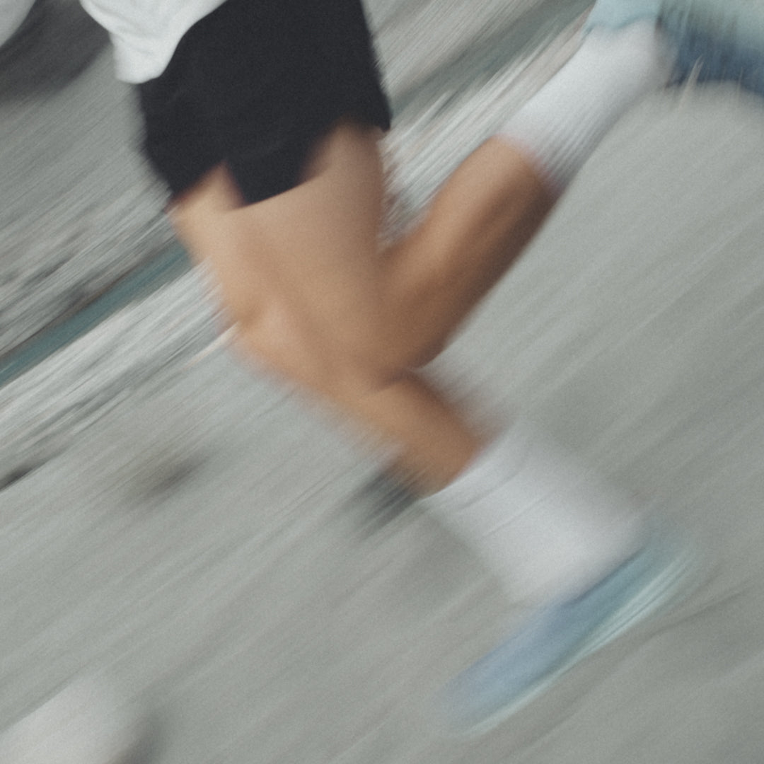 Verschwommene Nahaufnahme von Beinen eines laufenden Menschen, gekleidet in schwarze Shorts, weiße Socken und hellblaue Laufschuhe, auf einem grauen Hintergrund in Bewegung aufgenommen.