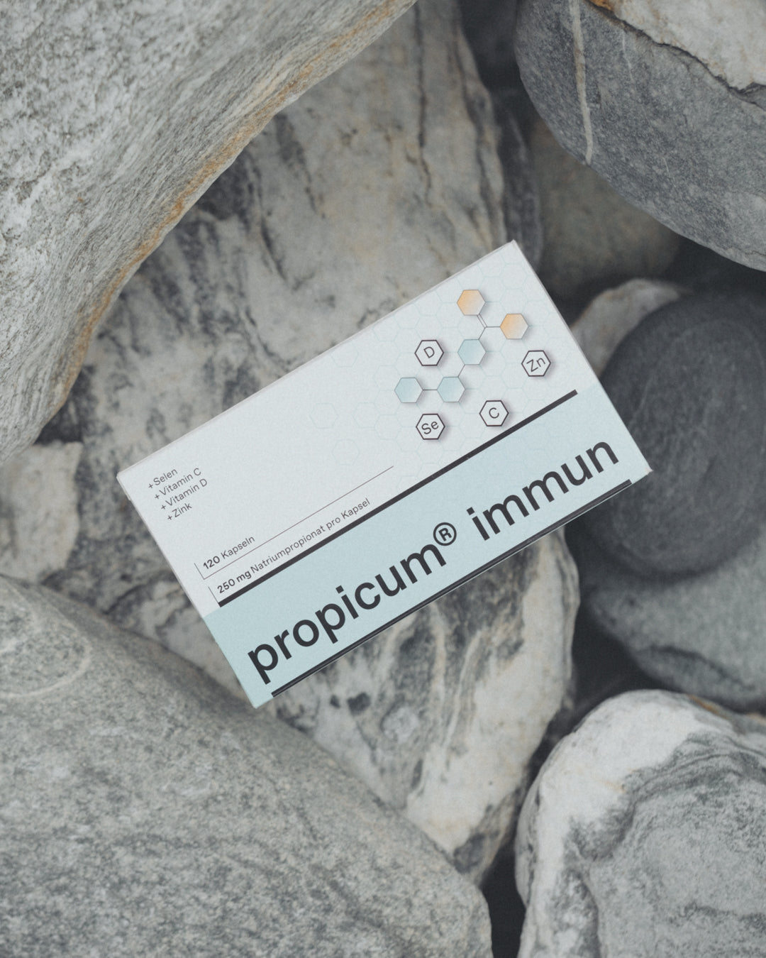 Verpackung von Propicum® Immun, platziert auf grauen Felsen im Freien. Das Design betont die Kombination aus Selen, Vitamin C, Vitamin D und Zink, die zur Stärkung des Immunsystems beitragen.