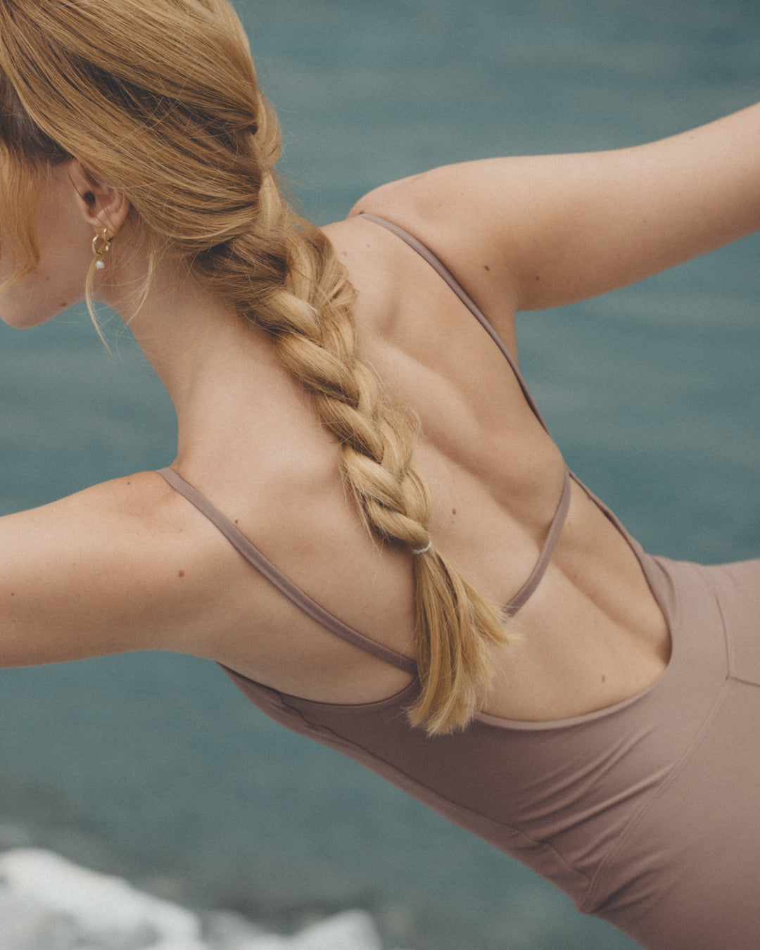 Frau mit blonder, geflochtener Frisur in einem beige-farbenen Yoga-Outfit, die eine Yoga-Pose am Flussufer praktiziert