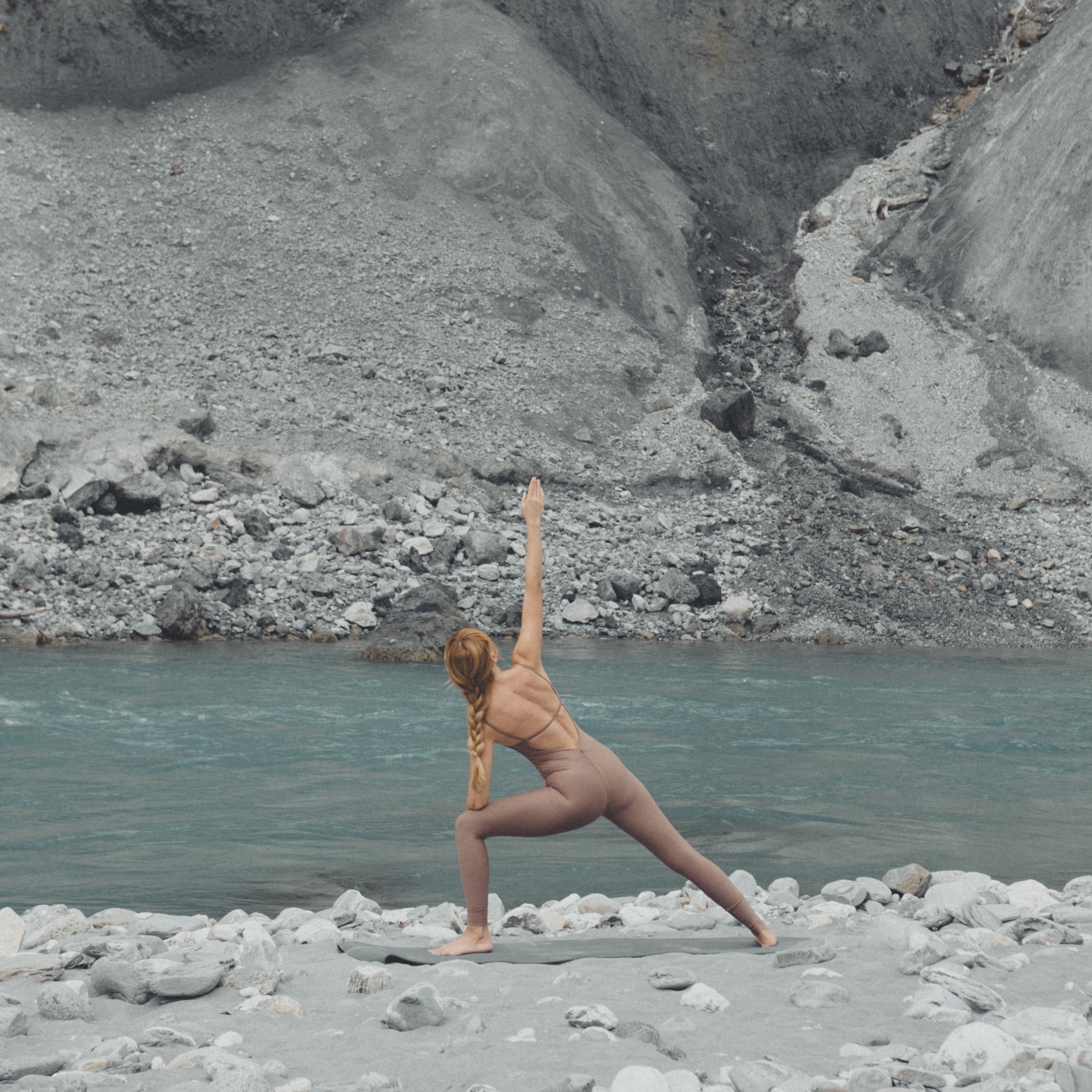 Frau in hautfarbenem Sportoutfit praktiziert eine Yoga-Pose am Ufer eines türkisfarbenen Gebirgsflusses vor einer kargen, steinigen Berglandschaft. Ihre Position vermittelt Stärke und Ruhe inmitten der naturbelassenen Umgebung.