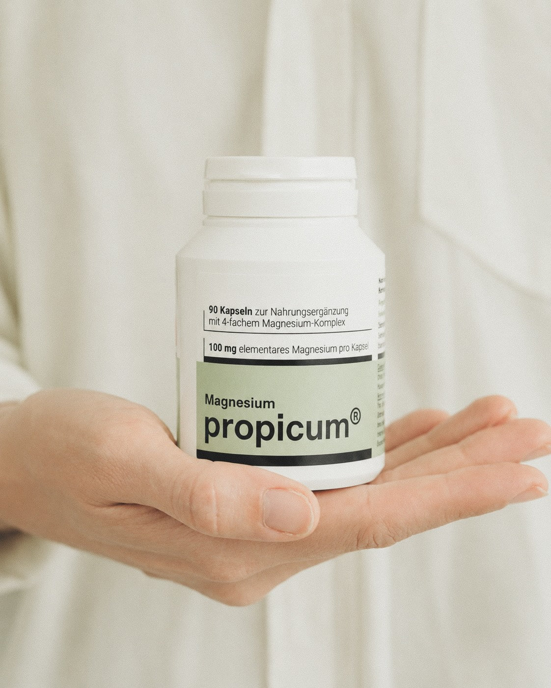 Eine grün weiße Plastikverpackung mit propicum Magnesium wird von einer Hand gehalten. Im Hintergrund sieht man eine Nahaufnahme eines weißes Hemdes