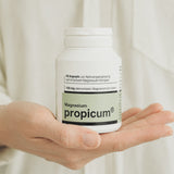 Eine grün weiße Plastikverpackung mit propicum Magnesium wird von einer Hand gehalten. Im Hintergrund sieht man eine Nahaufnahme eines weißes Hemdes