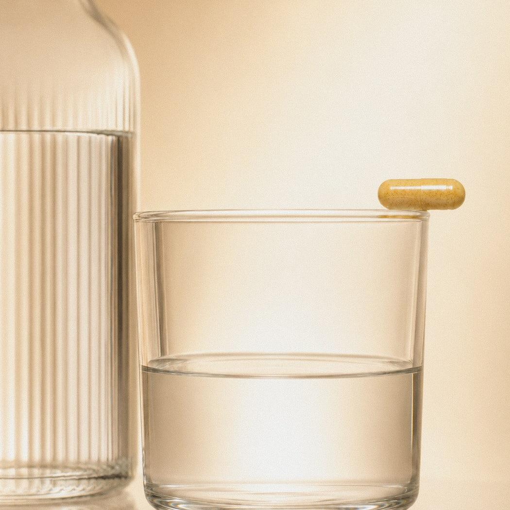 Eine kurkumafarbene Kapsel balanciert auf einem Glas Wasser neben einer Glaskaraffe