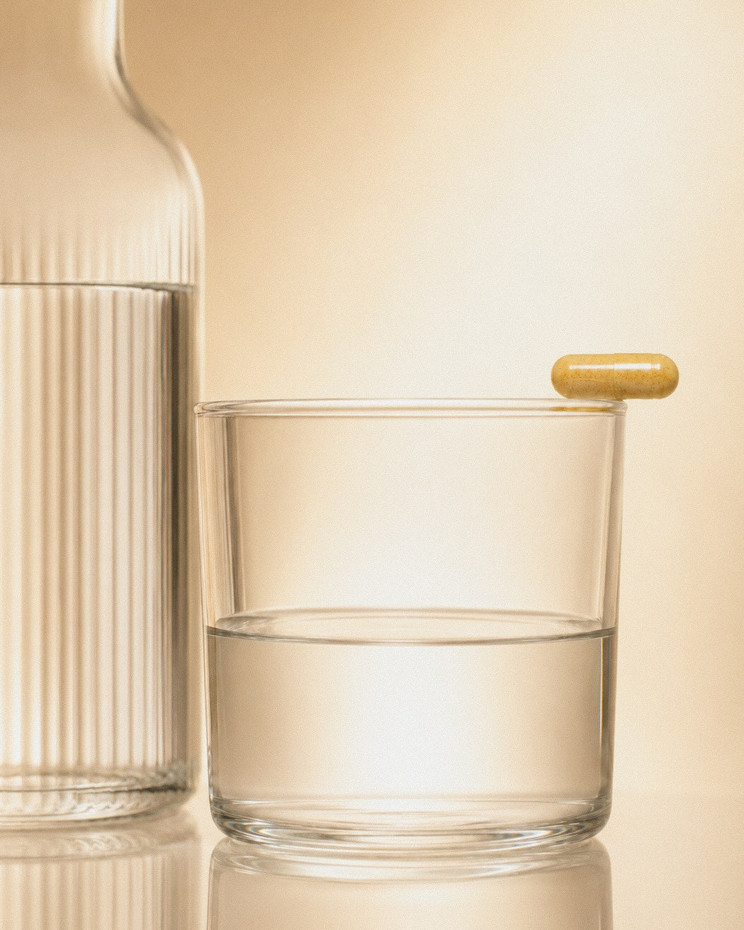 Eine kurkumafarbene Kapsel balanciert auf einem Glas Wasser neben einer Glaskaraffe
